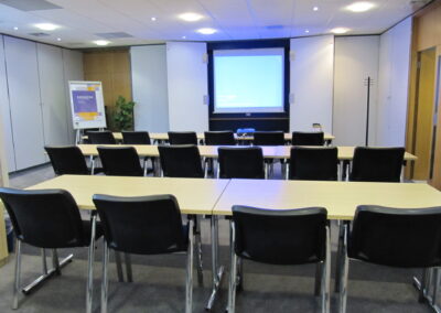 A conference room with three rows of tables and black chairs facing a blank projector screen, a projector on the front table, a flipchart to the left, and plain white walls.