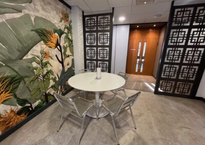 A small round table with four metal chairs is placed in a corner next to a large botanical mural. There is a clear water bottle on the table, and decorative black panels and a wooden door are visible in the background.