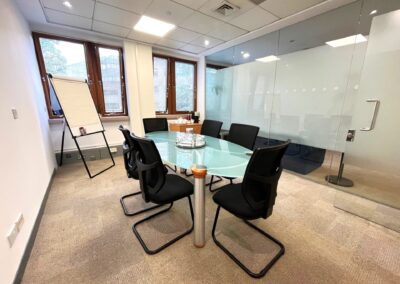 A modern conference room with a glass table surrounded by six black chairs, a flip chart, a glass wall, and three windows letting in natural light. There are glasses and water jugs on the table.