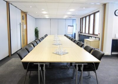 A conference room with a long, U-shaped table surrounded by black chairs, set with glass water jugs and glasses. The room has large windows, a flip chart, and a sideboard with cups and saucers.