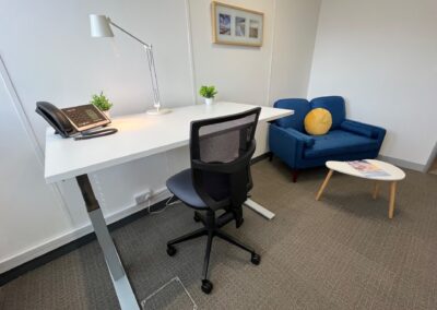 A modern office with a white desk, office chair, desk lamp, telephone, two small potted plants, a blue armchair with a yellow cushion, a small white table, and framed pictures on the wall.